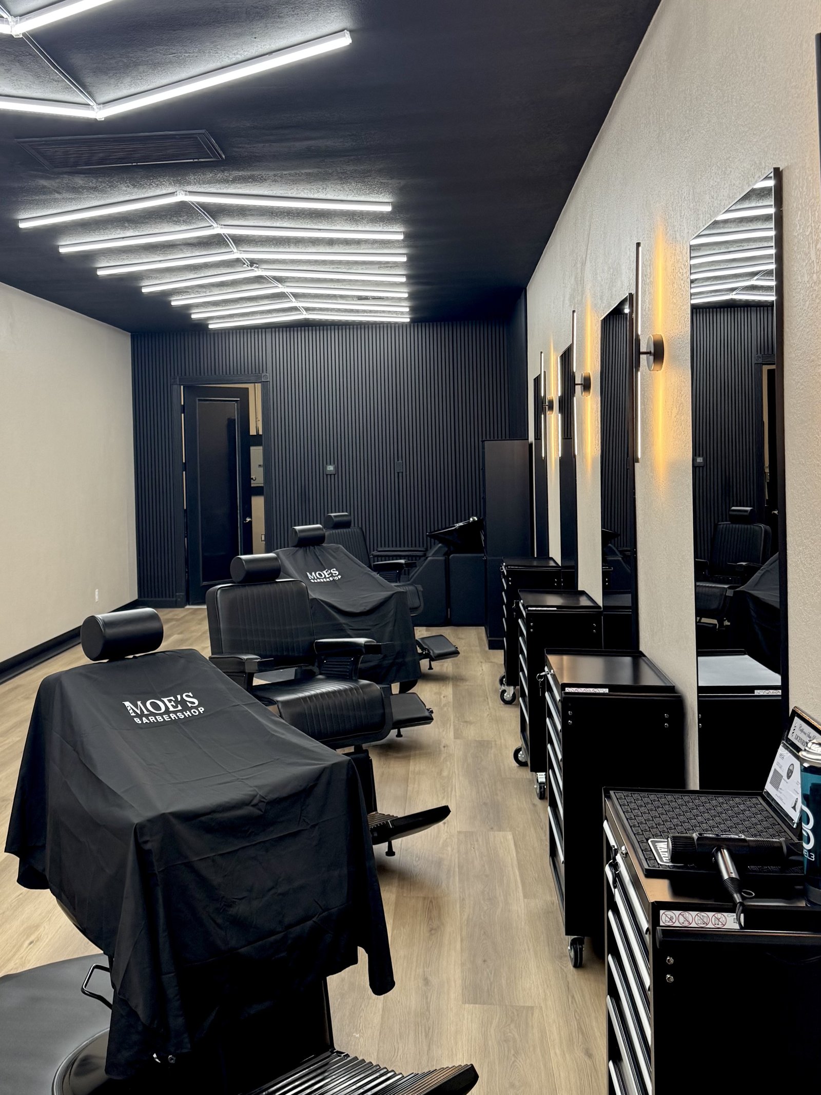 A row of black barber chairs under the angular ceiling lights at Moe's Barbershop